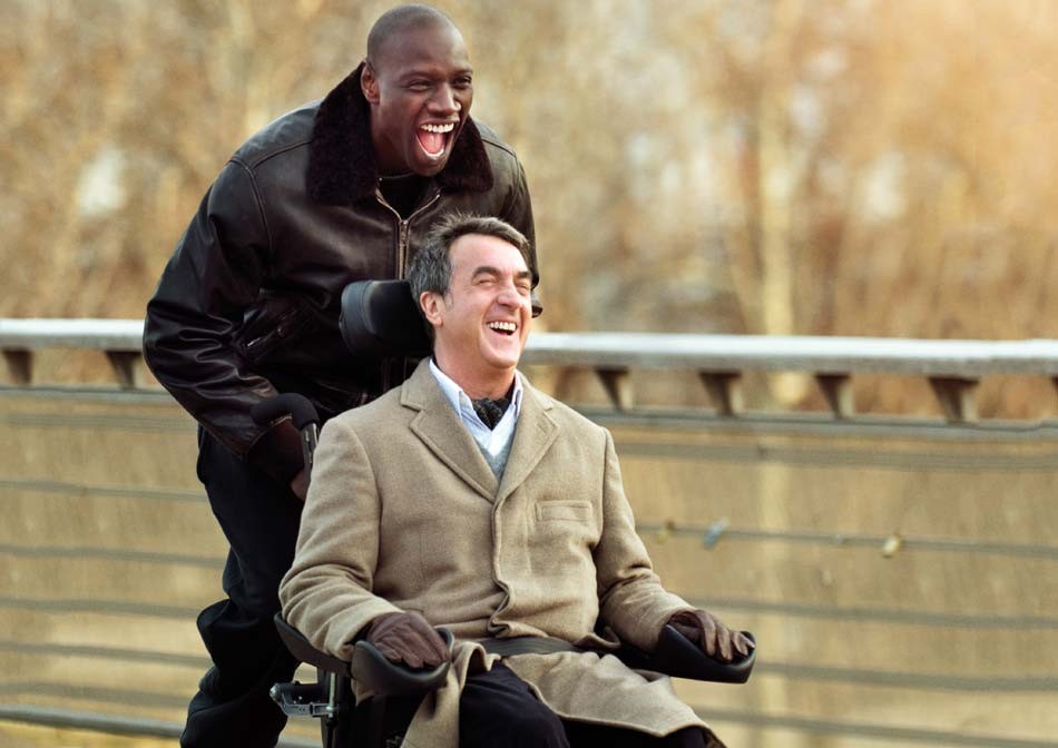 One man pushes the other on a wheelchair on a cold day, both of them happy